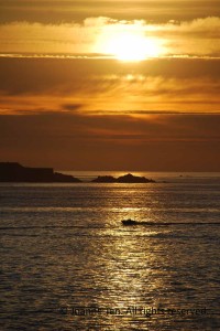A setting Sun through hazy clouds, a moving boat on golden water with sea rocks. Photographed on top of the old city wall overlooking the Atlantic Ocean, in Saint Malo, France, a popular beach town for summer vacation.