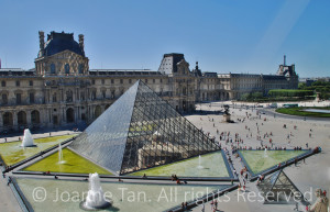Water fountains laying out in a pattern around the Glass Pyramid in the square outside the Louvre Palace. The picture was taken from inside of a glass of a tall building.