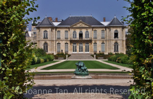Symmetrical French building's back door, window, and balcony overlook a rectangular lawn surrounded with a walkway and hedges carefully manicured. Rodin's statue of Infernal's blind father's cannibalism with his rear.