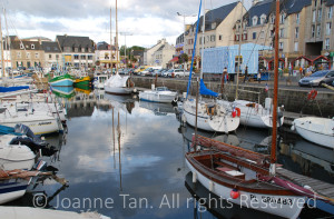 Stone buildings surround the port where boats dock, cloudy sky reflected in water. Picture taken in an old fishing village's downtown of Paimpol, Brittany, France.