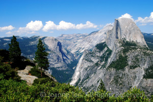Photographed on top of the Glacier Point of a bird's eye view of Half Dome in white clouds and blue sky, the Sierra Mountains, and the Yosemite Valley.