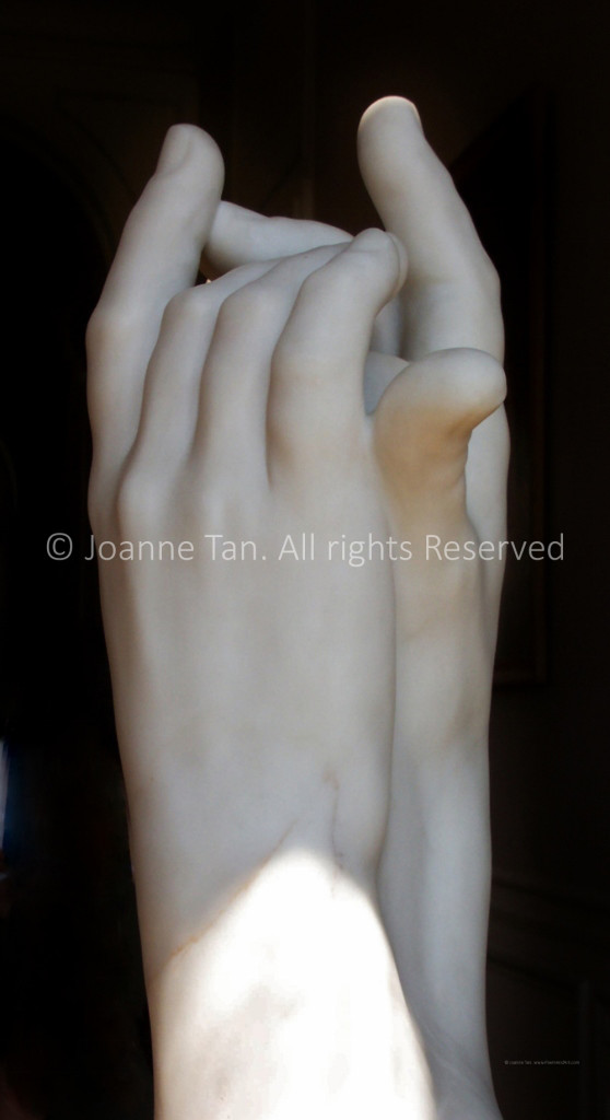 White marble statue of a pair of clasping gesture hands, pointing upwards, wrist and finger tip in partial sunlight, with a black background. Picture taken at the Rodin House, Paris, France.