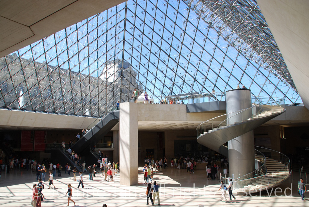An interior view towards the sky light ceiling & structure, with the old Louvre Palace & water shooting up from the fountain outside of the glass & metal pyramind. A modern structure with spiral staircase, free standing cube column, and sunlight on people walking inside.