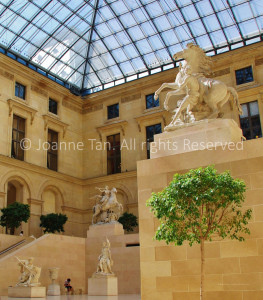Marble statues of warriors/soldiers with trumpet/bugle, weapons, and their horses, under glass ceiling's sky light above a 3-story-interior converted from exterior, inside the Louvre Museum, with indoor trees. Paris, France.