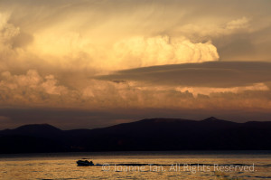 Burning clouds by the setting Sun over the dark mountain around the bright water ripples broken through by a moving motor boat.