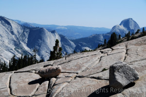 Two rocks in balance on a huge slap of granite with natural grooves and lines, in the blue background is the top of Half Dome and the granite domes and the Sierra Mountains in Yosemite, CA. An eagle flying against the granite mountain lined by pine forest.
