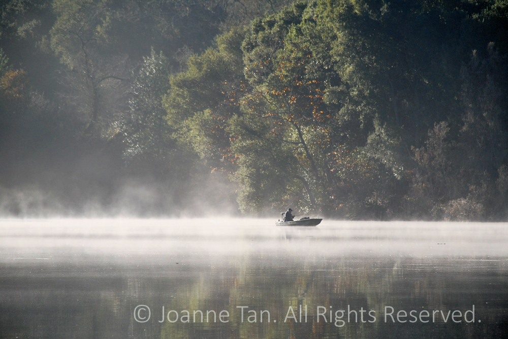 Morning fog over a steaming lake banked with early autumn foliage & trees, a lone fishing small boat afloat.